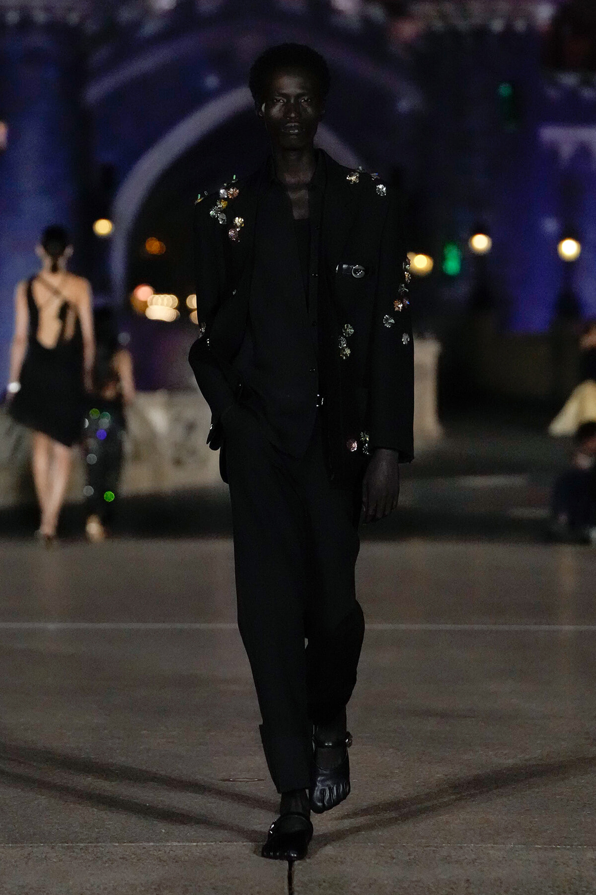 Model walking a night runway in a black embroidered suit with floral appliqués, moody blue lighting in background.