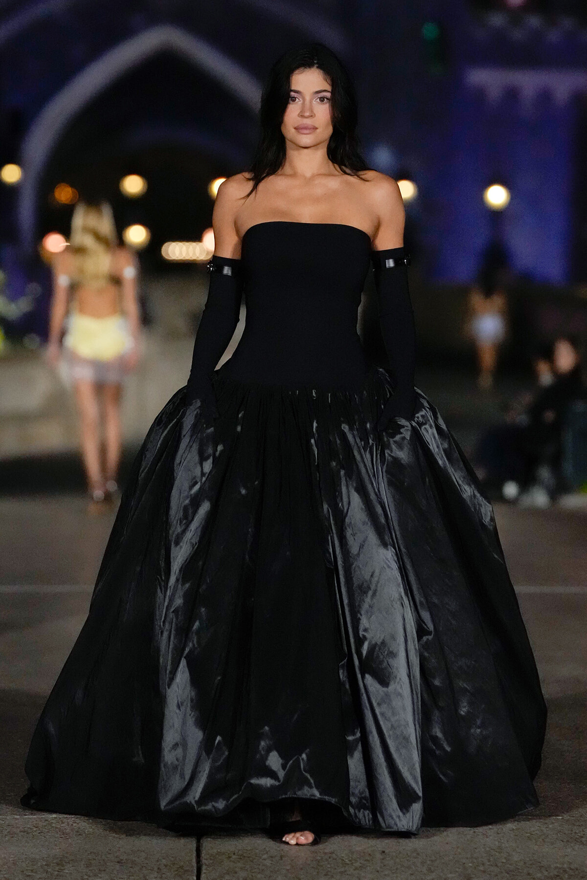 Model walking a strapless black ball gown with a full satin skirt on a runway at night.