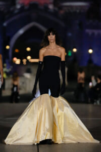 Model on a runway wearing a strapless black jumpsuit with dramatic cream satin mermaid skirt, gloves, and dark wavy hair.