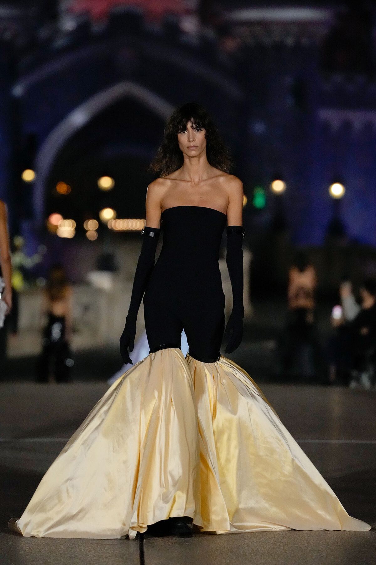 Model on a runway wearing a strapless black jumpsuit with dramatic cream satin mermaid skirt, gloves, and dark wavy hair.