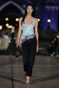 Model walking a runway at night in a pastel blue floral-embellished top and black wide-leg jeans, holding a small black clutch.