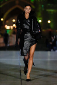 Model walking a nighttime fashion show in a shiny black textured coat with oversized collar, carrying a black handbag and wearing pointed heels.