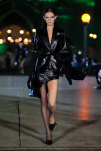 Model walking at night in a glossy black blazer-dress, thigh-high slit, black gloves, and carrying a large black bag.