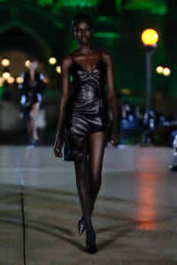 Model in a black metallic mini dress and pointed heels walking on a nighttime outdoor runway with blurred lights in the background.