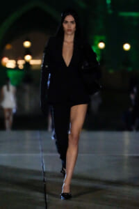 Woman walking at night in a black plunging blazer-dress with one leg visible, carrying a large black bag; green street lighting in the background.