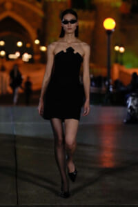Female model in a black strapless mini dress and sunglasses walking at night on a city street.