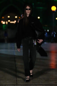 Person in all-black outfit wearing a hoodie and sunglasses, walking at night with a black bag over the shoulder.
