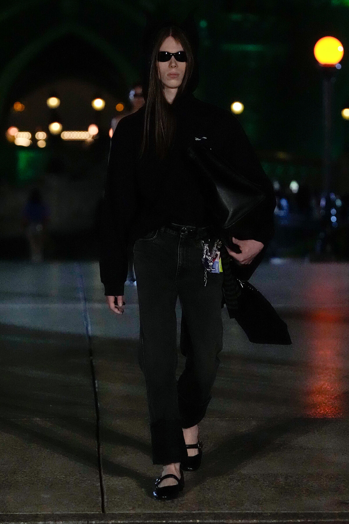 Person in all-black outfit wearing a hoodie and sunglasses, walking at night with a black bag over the shoulder.