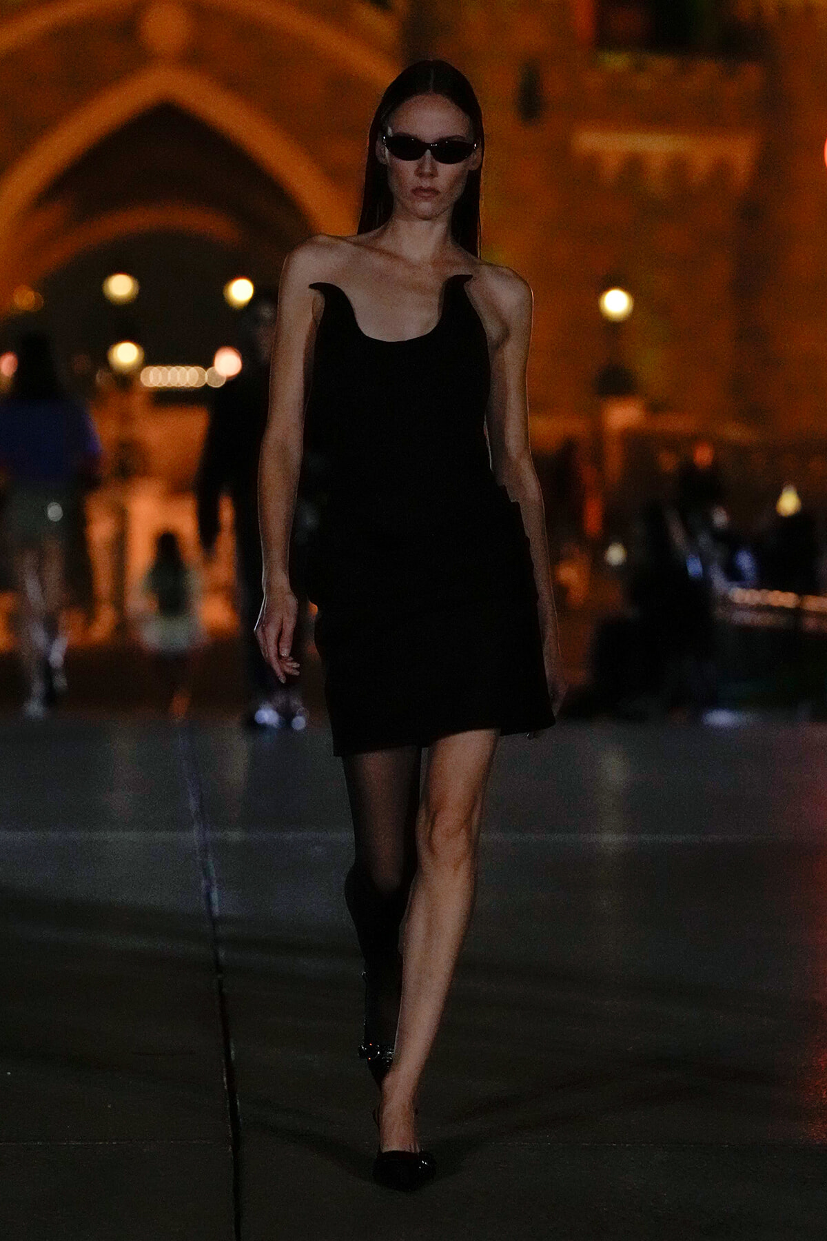 Model in a black sleeveless mini dress and sunglasses walks along a dimly lit city street at night.