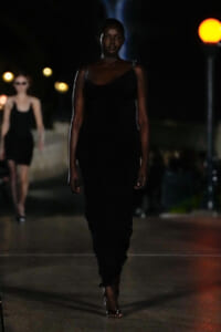 Model walking down a dimly lit street runway in a black evening gown, foreground focus on her serious expression and silhouette.