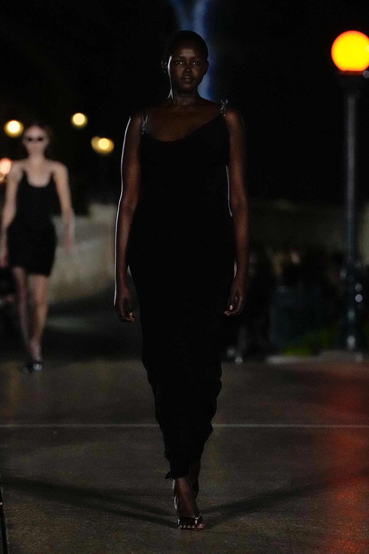 Model walking down a dimly lit street runway in a black evening gown, foreground focus on her serious expression and silhouette.