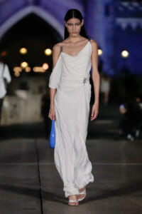 Model walking a white draped gown on a runway at night, carrying a bright blue clutch.