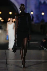 Dark-skinned model walks a black, asymmetric mini dress down a nighttime runway, with out-of-focus background and lights behind.