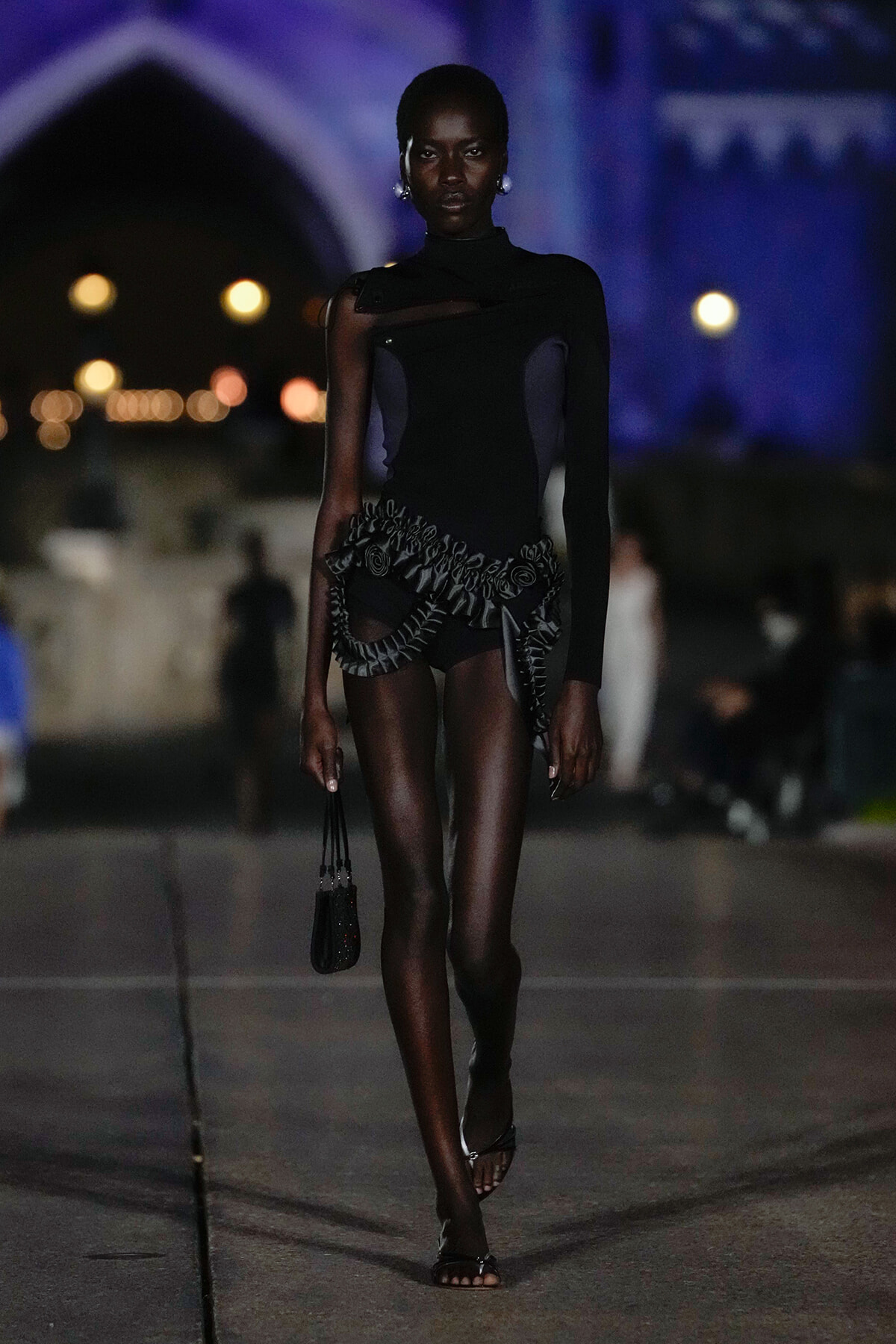 Model walking runway at night in a black, cut-out bodysuit with ruffled harness and small black bag.