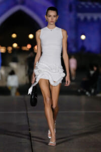 Model walking a fashion runway in a sleeveless white mini dress with sculpted ruffle details at the hem, carrying a small black patent bag.