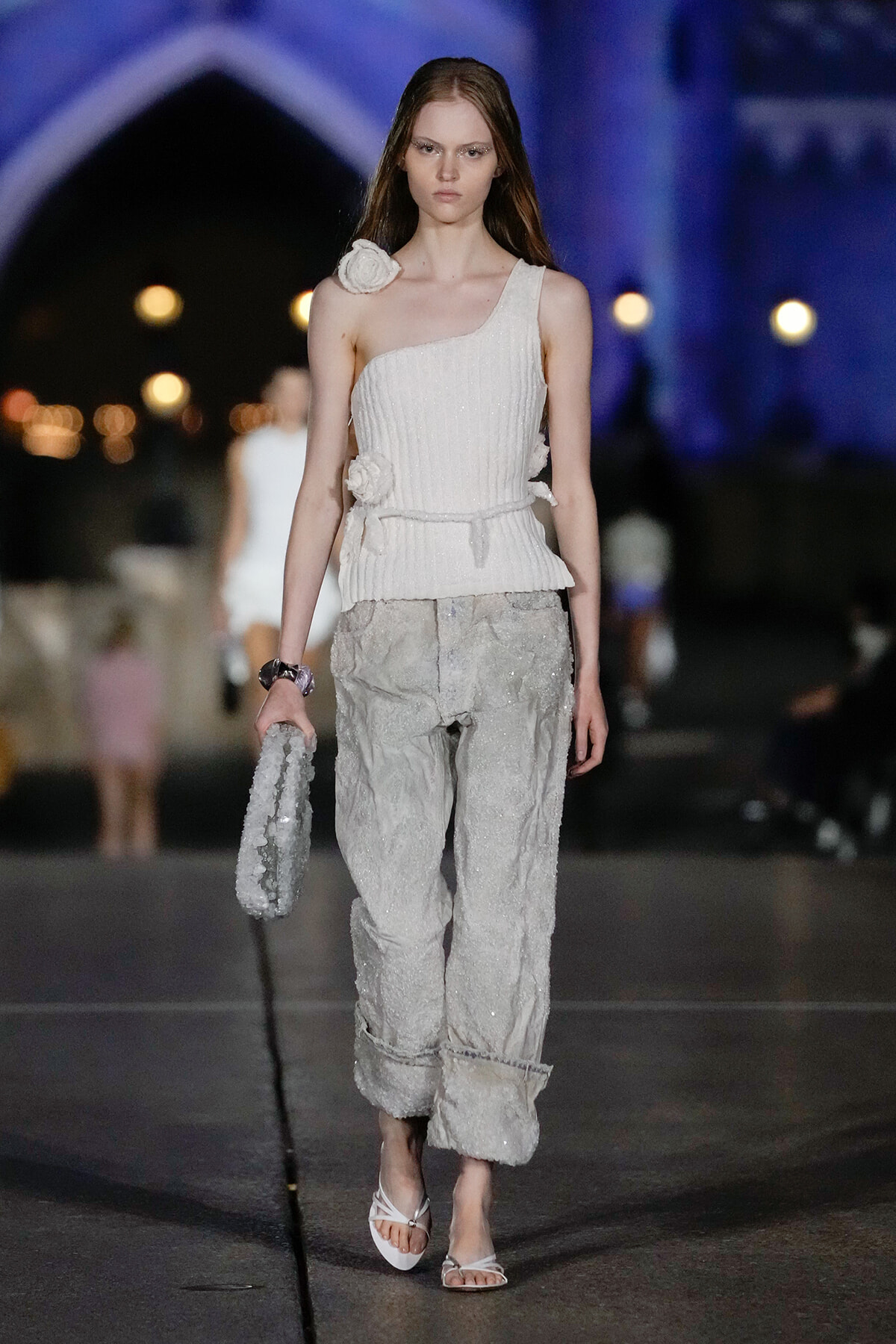 Model walks a runway in an all-white one-shoulder knit top with flower embellishments and textured light-gray pants, holding a beaded clutch.