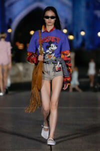 Model walking on a runway at night in a blue Disney T-shirt, shorts, sunglasses, and fringe brown bag; urban backdrop with bokeh lights.