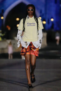 Model walks a fashion runway at night in a pale yellow tee with 'Coperni princess' text, white embroidered sleeves, tropical orange shorts, and black sandals with sunglasses.