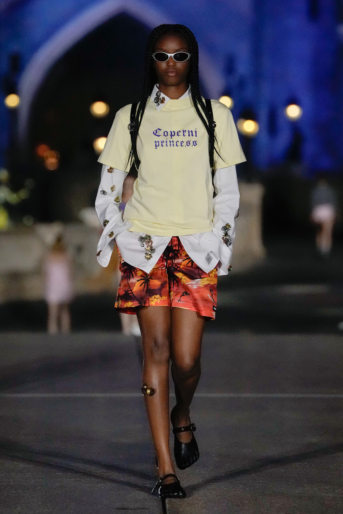 Model walks a fashion runway at night in a pale yellow tee with 'Coperni princess' text, white embroidered sleeves, tropical orange shorts, and black sandals with sunglasses.