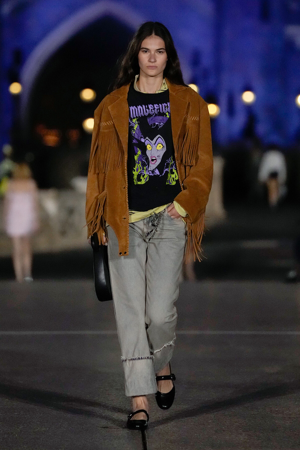 Model walks runway in a tan fringed suede jacket over a graphic black tee, light gray cuffed jeans, and black Mary Jane shoes at night.