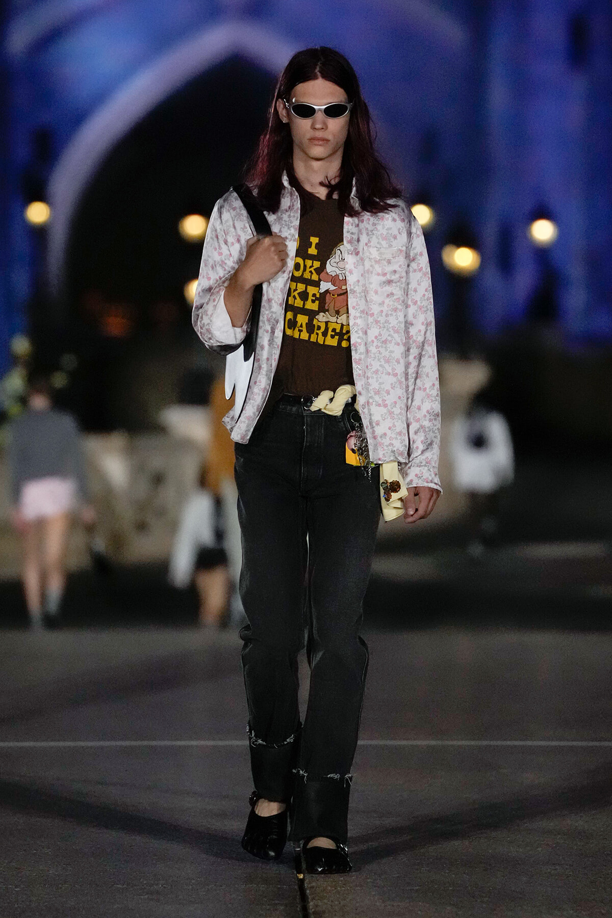 Model walking a runway at night in a white floral jacket, brown graphic tee, black jeans, and white sunglasses.