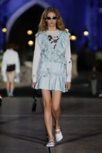 Model walking a runway in a pale blue, ruffled vest over a white Mickey Mouse tee and matching mini dress with white shoes and sunglasses.