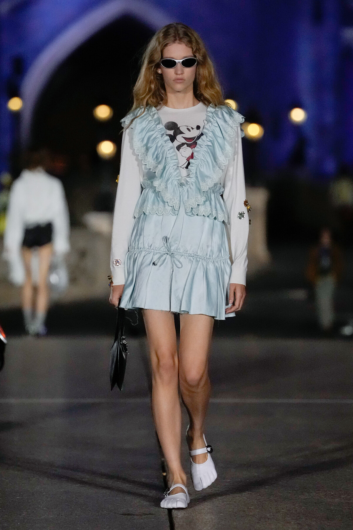 Model walking a runway in a pale blue, ruffled vest over a white Mickey Mouse tee and matching mini dress with white shoes and sunglasses.