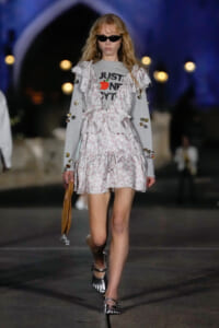 Model walking on a runway wearing a light floral dress over a gray sweatshirt with 'JUST ONE BYT' text, sunglasses, and metallic sandals, holding a tan handbag.