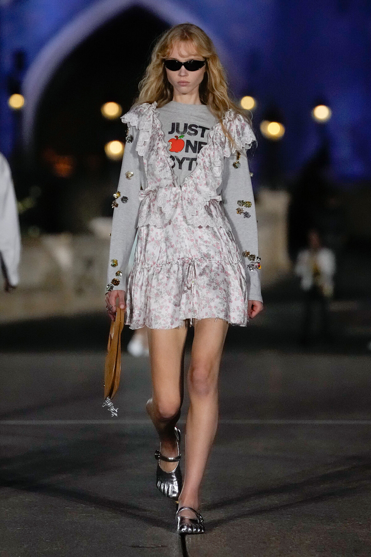 Model walking on a runway wearing a light floral dress over a gray sweatshirt with 'JUST ONE BYT' text, sunglasses, and metallic sandals, holding a tan handbag.
