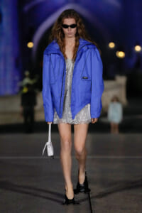 Model walking a runway in a bright blue oversized jacket over a silver sequin mini dress, wearing black sunglasses and carrying a small white handbag with black heels.