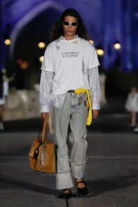Model on a runway wearing a white oversized T-shirt over a sequined long-sleeve shirt, light gray jeans, sandals, and sunglasses, carrying a tan tote bag with accessories.