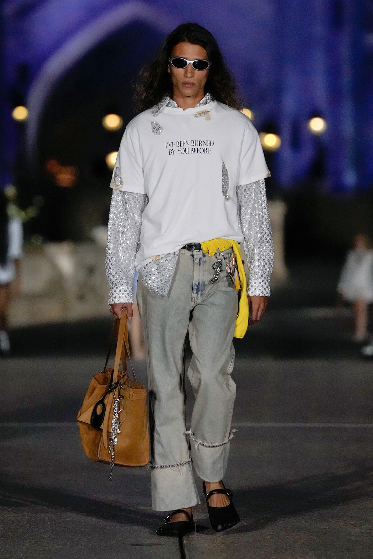 Model on a runway wearing a white oversized T-shirt over a sequined long-sleeve shirt, light gray jeans, sandals, and sunglasses, carrying a tan tote bag with accessories.