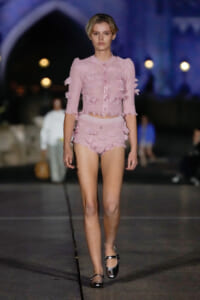Model wearing a pale pink, ruffled two-piece outfit walking a fashion show runway at night.