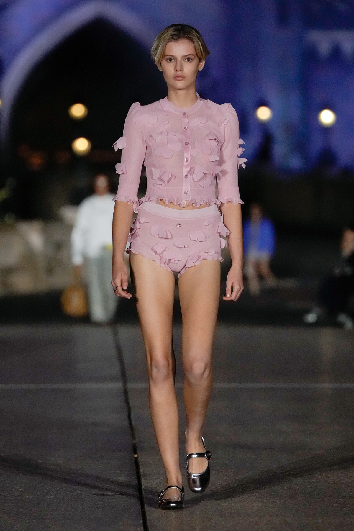 Model wearing a pale pink, ruffled two-piece outfit walking a fashion show runway at night.