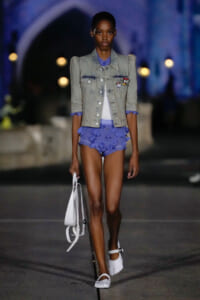 Model walks a runway wearing a light denim jacket over a purple ruffled shorts set, carrying a white handbag and wearing white sandals in a dimly lit scene.