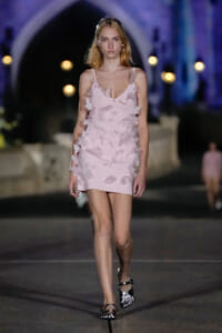 Model walking a night runway in a pale pink mini dress with 3D petal embellishments against a blue-lit backdrop.
