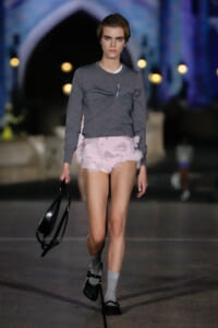 Model walking a runway at night, wearing a gray knit top and pale pink ruffled shorts, black bag in hand.