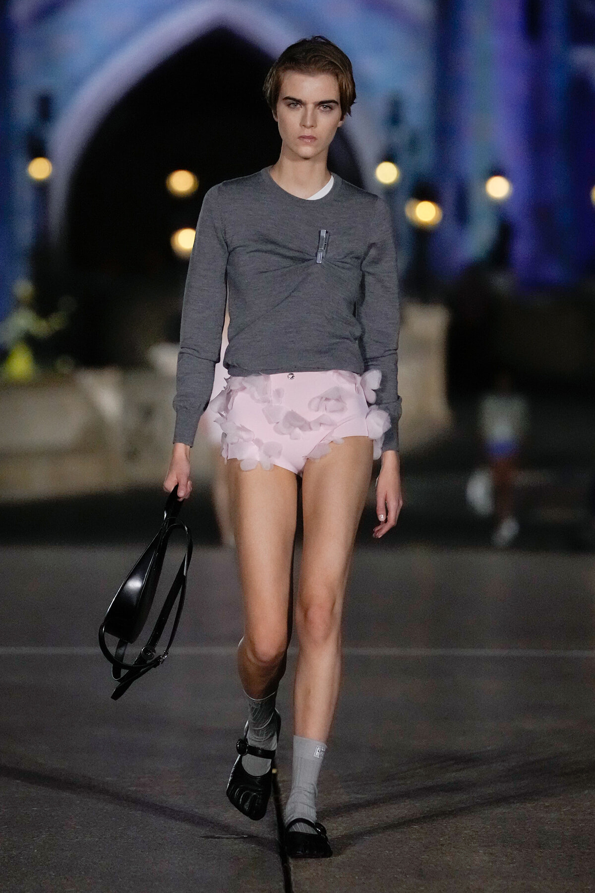 Model walking a runway at night, wearing a gray knit top and pale pink ruffled shorts, black bag in hand.