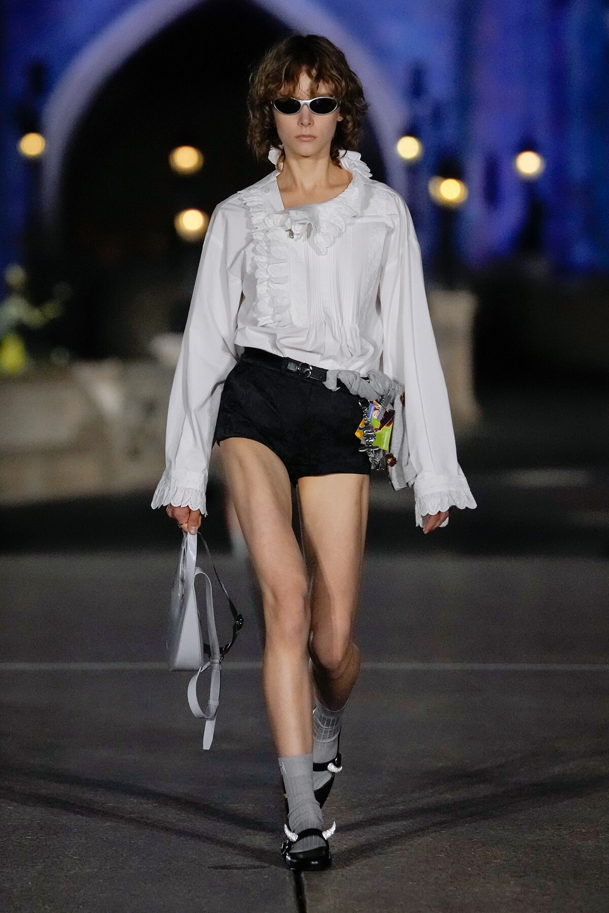 Model walking a runway in a white ruffled blouse and black shorts, wearing sunglasses and carrying a light bag and accessories.