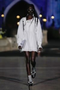 Model walking a night runway in a white ruffled dress with lace trim, white sunglasses, and braided hair, carrying a booklet
