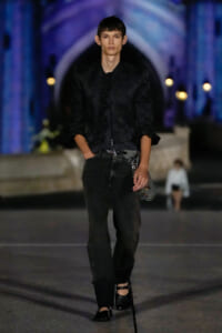 Male model walking a dimly lit runway in an all-black outfit: textured jacket, dark jeans, and black shoes with a small bag at the hip.