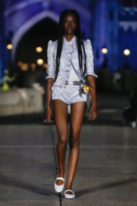 Model walking on runway in light gray floral suit with ruffled front and matching shorts, white belt and Mary Jane shoes under blue-lit venue.