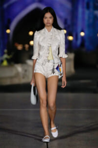 Female model walks a white satin suit with ruffled blouse and matching shorts on a fashion runway in a dimly lit venue.