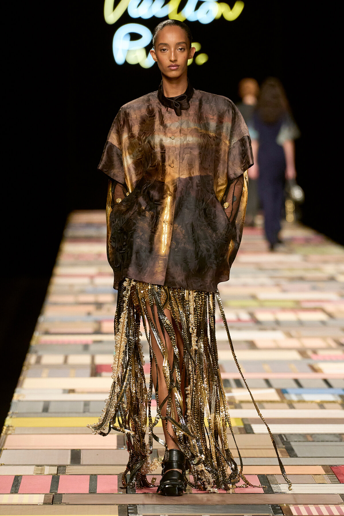 Model walks a fashion runway in a metallic bronze sheer tunic with a fringe, bejeweled skirt cascading to the floor.