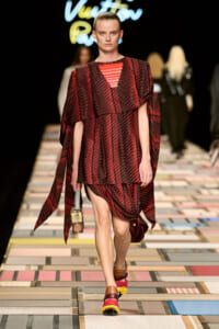 Model on a runway wearing a red plaid cape-style dress with an orange pyramidal neckline, carrying a small handbag and wearing chunky multicolor sandals.