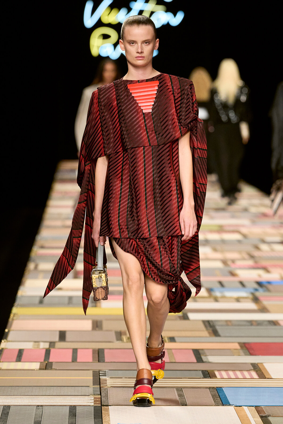 Model on a runway wearing a red plaid cape-style dress with an orange pyramidal neckline, carrying a small handbag and wearing chunky multicolor sandals.