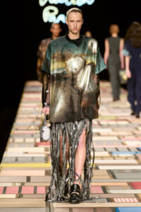 Model on a runway wearing an oversized metallic landscape-print top with sheer sleeves and a long fringe-bead skirt; carrying a small metallic box purse.