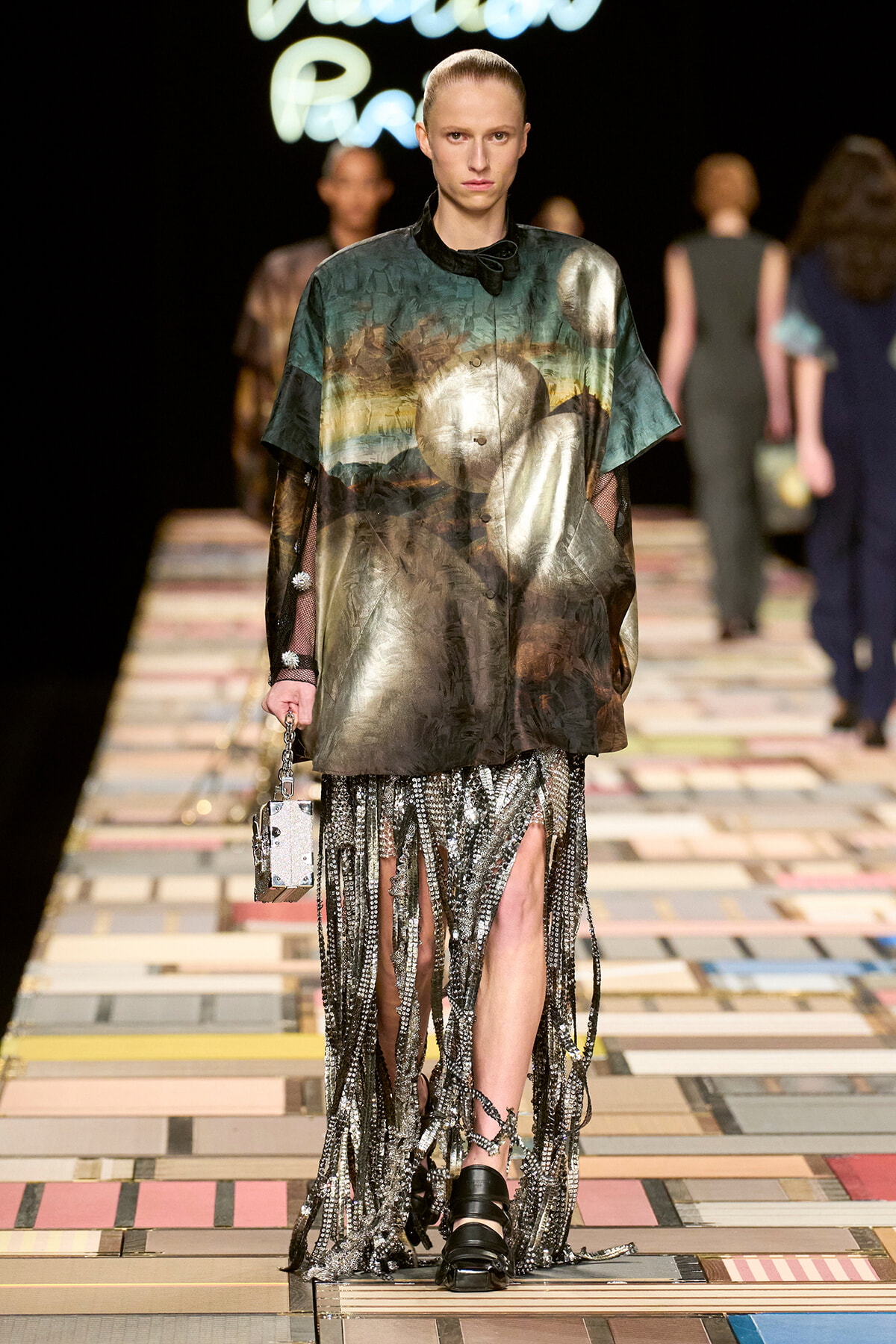 Model on a runway wearing an oversized metallic landscape-print top with sheer sleeves and a long fringe-bead skirt; carrying a small metallic box purse.
