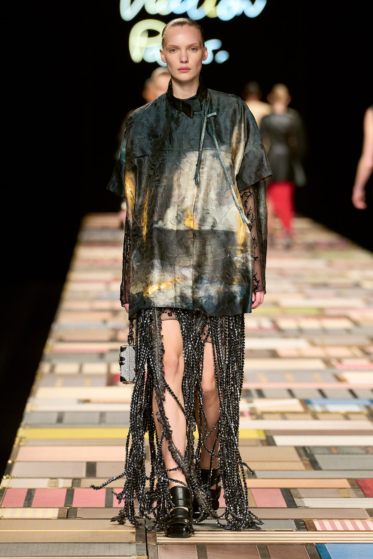 Model on a runway wearing an oversized abstract-print top and a floor-length beaded fringe skirt, walking on a colorful tiled catwalk.