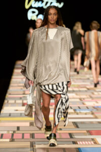 Model walks the runway in a loose metallic silver striped poncho with a tied striped wrap skirt and a light handbag.